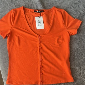 Orange v-ringad topp från Bik Bok - Fräsch orange topp från Bik Bok i storlek S. Modellen har v-ringning, korta ärmar och knappar längs hela framsidan. Tunn och mjuk bomullskvalitet som känns skön mot huden. Perfekt för dig som gillar färgstarka och stilrena plagg. Den är aldrig använd helt ny.