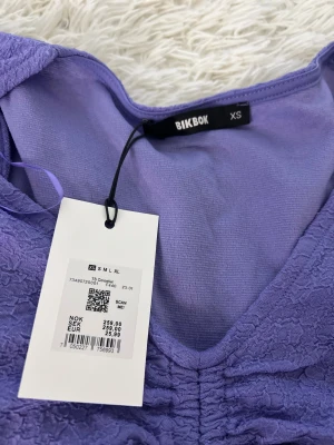 Lila omlottopp med snörning från Bikbok - Supersöt lila topp från Bikbok i storlek XS. Modellen har omlottdetalj och snörning framtill som ger en snygg rynkad effekt. Puffiga korta ärmar och v-ringad hals. Materialet är mjukt och har en lätt struktur som känns skön mot huden.