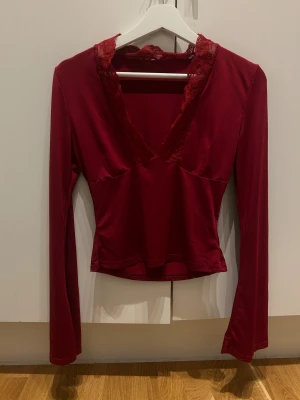 Vinröd långärmad topp med spets - Säljer en vinröd långärmad topp med djup v-ringning och dekorativ spets runt halsen. Toppen har en figurnära passform och är tillverkad i ett mjukt, glansigt syntetmaterial som känns skönt mot huden. Perfekt för dig som gillar romantiska detaljer och snygg siluett.