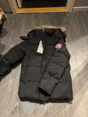 Canada goose - Säljer en svart dunjacka från Canada Goose med huva och avtagbar päls . Jackan har klassisk patch på ärmen, stora fickor och dragkedja med knappar. Perfekt för kalla vinterdagar och riktigt snygg streetstil.