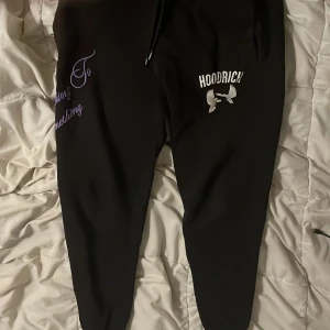 Svarta Hoodrich mjukisbyxor strl M - Svarta mjukisbyxor från Hoodrich med tryckt logga och textdetaljer i vitt och lila. Byxorna har snörning i midjan och ribbade muddar vid bensluten. Skön passform och mjukt material, perfekt för chill eller streetwear.