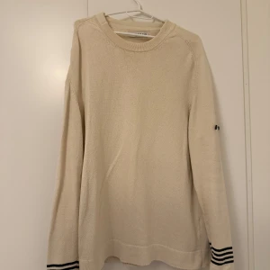 Beige stickad tröja Stone Island - Snygg beige stickad tröja från Stone Island med svarta knappar på ena ärmen och svarta ränder vid ärmsluten. Tröjan har rund hals och lång ärm, perfekt för dig som gillar clean och stilren design. Liten i storlek mer som XL eller större L.