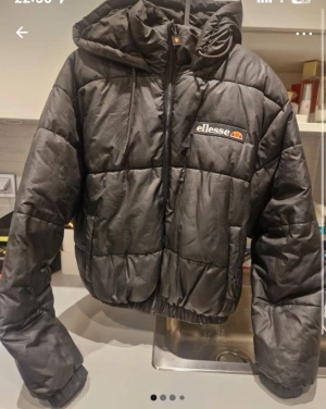 Svart pufferjacka från Ellesse - Svart pufferjacka från Ellesse med huva och dragkedja framtill. Jackan har quiltad design, elastiska muddar och Ellesse-logga på bröstet. Perfekt för kalla dagar och har en loose passform som ger en chill vibe.