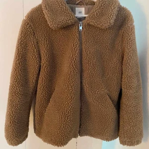 Beige teddyjacka från H&M M/L i storlek  - Mysig teddyjacka från H&M i beige färg med fluffig och mjuk yta. Jackan har en klassisk krage, dragkedja framtill och långärmade ärmar. Perfekt för att hålla värmen under kyliga dagar och ger en chill vibe till din outfit. Storleken är M/L passar båda. Helt oanvänd endast provad så är i nyskick. 