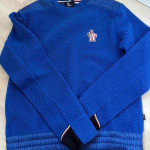 Blå fleece-tröja från Moncler Grenoble - Säljer en blå fleece-tröja från Moncler Grenoble i storlek M. Tröjan har quiltade detaljer på axlar och nederkant, samt ett broderat Moncler-märke på bröstet. Långärmad och med ribbade muddar i svart med röd-vit rand. Perfekt för kyliga dagar.