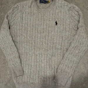 Grå kabelstickad tröja Polo Ralph Lauren - Snygg grå kabelstickad tröja från Polo Ralph Lauren med klassisk rund hals och broderad logga på bröstet. Tröjan har långa ärmar och ribbade muddar vid ärmslut och nederkant. Perfekt för dig som gillar stilren och tidlös design.