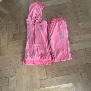 Juicy Couture rosa hoodie med dragkedja - Rosa hoodie från Juicy Couture i mjuk velour med silverfärgat logotryck på bröstet och ryggen. Tröjan har huva, långa ärmar, fickor fram och dragkedja. Perfekt för chill eller att styla med matchande byxor för en ikonisk Y2K-look.