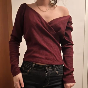Vinröd omlottopp med offshoulder - Snygg vinröd omlottopp med offshoulder och lång ärm. Toppen har en djup v-ringning och är tillverkad i ett ribbat material som ger en cool struktur. Passformen är normal och toppen sitter snyggt över axlarna. Priset går att buda på🥰