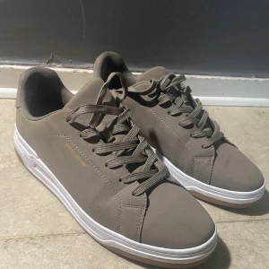 Grå sneakers från Bruno Marc - Snygga grå sneakers från Bruno Marc med stilren design. Skorna har rund tå, platt vit sula och matchande grå snörning. Ovandelen är i syntetmaterial med diskret logga i guld på sidan. Perfekta för en clean och modern look.