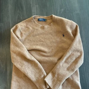Kashmir tröja från Polo Ralph Lauren - Snygg beige tröja från Polo Ralph Lauren i Kashmir. Klassisk rund hals och ribbade muddar vid ärmslut och nederkant. Liten mörkblå logga broderad på bröstet. Perfekt för dig som gillar stilrena och tidlösa plagg. Finns en liten defekt som knappt syns (bild 2)