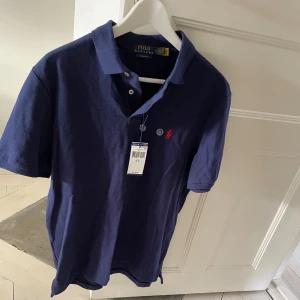 Blå pikétröja från Polo Ralph Lauren - Helt ny och oanvänd snygg mörkblå pikétröja från Polo Ralph Lauren i klassisk passform. Tröjan har korta ärmar, krage med två knappar och den ikoniska röda broderade loggan på bröstet. Tillverkad i mjuk bomull som känns skön mot huden.