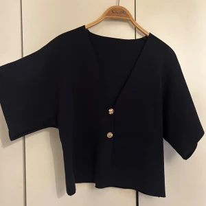 Svart blus med guldknappar - Säljer en mörkblå blus med korta ärmar och djup v-ringning. Blusen har två snygga guldknappar framtill och är tillverkad i ett ribbat material som ger en cool struktur. toppen är perfekt till fest eller vardags och för dig som gillar stilrena detaljer💙