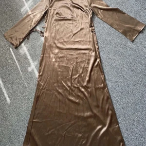 Brun långklänning i satin - Snygg brun långklänning i glansigt satinliknande material med långa ärmar och rund halsringning. Klänningen har smala knytband i midjan för en justerbar passform. Perfekt för dig som gillar en elegant och stilren look.