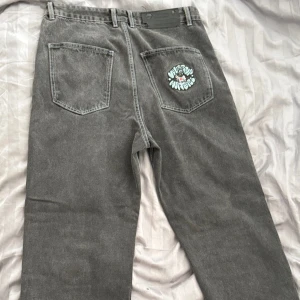 Grå jeans med broderi från Wasted - Grå jeans med raka ben och coola broderier av en tecknad figur på både bakficka och framficka. Jeansen har klassisk fyraficksdesign, kontrastsömmar och är tillverkade i kraftigt denimtyg. Perfekt för dig som vill sticka ut med streetwear-vibe.
