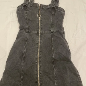 Svart jeansklänning med dragkedja - Cool svart jeansklänning med justerbara axelband och en snygg silverfärgad dragkedja framtill. Klänningen är kort och har en figurnära passform som framhäver siluetten. Perfekt för dig som gillar en edgy och trendig stil.