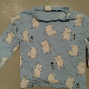 Blå Mumin långärmad tröja Lindex - Supersöt ljusblå långärmad tröja från Lindex med Mumin-figurer över hela plagget. Tröjan har tryckknappar vid halsen för enkel påklädning och är gjord i mjuk bomull. Perfekt för dig som gillar Mumin och vill ha något bekvämt och gulligt.