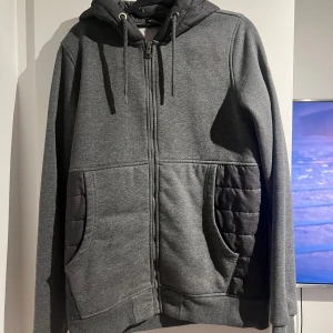 Grå Moncler hoodie med quiltade detaljer - Säljer en grå hoodie från Moncler med quiltade partier på fickor, rygg och huva. Tröjan har dragkedja framtill, snörning i huvan och broderad Moncler-logga på ärmen. Perfekt för kyliga dagar och riktigt snygg streetstil.
