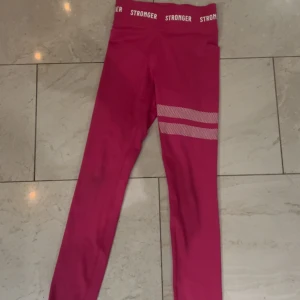 Rosa leggings från Stronger - Snygga rosa leggings från Stronger med vit logga i midjan och två vita prickiga ränder på ena benet. De har hög midja och är tillverkade i ett stretchigt syntetmaterial som passar perfekt för träning. Superskön passform och cool design.
