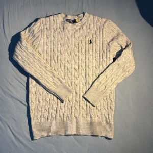 Ljusgrå stickad tröja Polo Ralph Lauren - Säljer en ljusgrå stickad tröja från Polo Ralph Lauren med klassiskt kabelstickat mönster och broderad logga på bröstet. Tröjan har rund hals, ribbade muddar och långärmad passform. Perfekt för dig som gillar stilren och tidlös design.