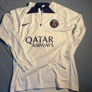 PSG långärmad vit fotbollströja Nike - Paris Saint-Germain långärmad fotbollströja från Nike i vit med mörkblå detaljer vid kragen. PSG-logga på bröstet och Qatar Airways tryck på framsidan. Tillverkad i lätt och ventilerande material, perfekt för match eller träning.