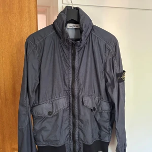 Grå vindjacka från Stone Island - Snygg grå vindjacka från Stone Island med klassisk badge på ärmen. Jackan har hög krage, två stora fickor med knappar framtill och dragkedja. Materialet är lätt och känns skönt mot kroppen, perfekt för dig som gillar streetwear och stilren design.