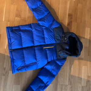 Blå dunjacka från Ralph Lauren S -  Jackan är köpt för 5500 kr gillade inte hur den satt så Säljer en fet blå dunjacka från Ralph Lauren, modell El Cap Jacket. Jackan har huva, svarta detaljer upptill och cool gul dragkedja på bröstfickan. Broderad logga på framsidan och två sidofickor med dragkedja. Perfekt för kalla dagar. Kan gå t lägre pris vid snabb affär
