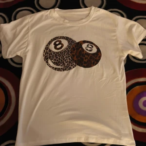 Vit t-shirt med leopardmönster tryck - Vit t-shirt med coolt tryck av två biljardbollar med leopardmönster på bröstet. Klassisk rund hals och korta ärmar. Materialet känns mjukt och är troligtvis bomull, perfekt för dig som gillar unika och djuriska detaljer.