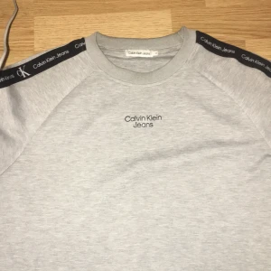 Grå sweatshirt från Calvin Klein Jeans - Snygg grå sweatshirt från Calvin Klein Jeans med svart logoband längs axlar och ärmar. Tröjan har rund hals och tryckt logga på bröstet. Perfekt för dig som gillar stilrena och klassiska plagg med en modern touch.