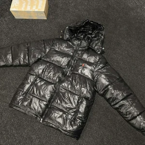 Svart pufferjacka från Polo Ralph Lauren - Säljer en svart pufferjacka från Polo Ralph Lauren med glansig finish och huva. Jackan har två framfickor med dragkedja, elastiska muddar och klassisk röd Polo-logga på bröstet. Perfekt för kalla dagar och har en riktigt schysst streetstil.