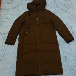 Svart lång pufferjacka  - Svart lång pufferjacka från MaxRita i storlek XL. Jackan har huva, två snedställda fickor med dragkedja och hel dragkedja framtill. Den quiltade designen ger extra värme och jackan har en clean look som passar perfekt när det är kallt ute.