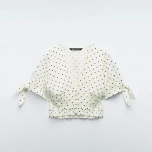 Vit prickig blus från Zara - Superfin vit blus från Zara med svarta prickar, v-ringad och croppad passform. Blusen har korta ärmar med knyt vid ärmslut och en söt volangkant nedtill. Perfekt för dig som gillar romantisk och trendig stil.