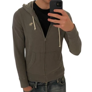 Grå hoodie från Abercrombie & Fitch | M - Säljer en grå hoodie från Abercrombie & Fitch i storlek M. Tröjan har dragkedja, huva med vita snören och broderad älg-logga på bröstet. Mjuk bomullskänsla och känguruficka framtill. Perfekt för chill dagar.