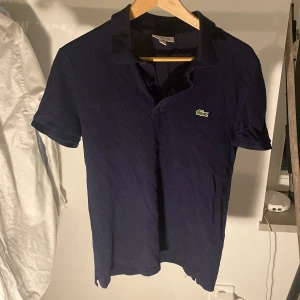 Mörkblå pikétröja från Lacoste - Snygg mörkblå pikétröja från Lacoste i slim fit-modell. Tröjan har klassisk krage, korta ärmar och den ikoniska gröna krokodilloggan broderad på bröstet. Tillverkad i bomull för en skön känsla och stilren look. 199 kr