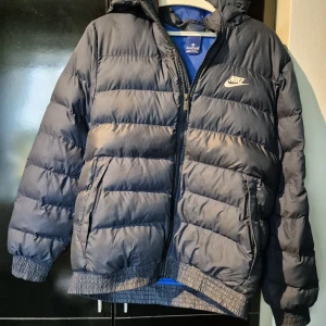 Svart pufferjacka från Nike XL - Snygg svart pufferjacka från Nike med huva och dragkedja. Jackan har ribbade muddar, två sidofickor och klassisk Nike-logga på bröstet. Perfekt för kalla dagar och riktigt skön att bära.