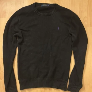 Svart stickad tröja från Polo Ralph Lauren - Svart stickad tröja från Polo Ralph Lauren med lila broderad logga på bröstet. Rund halsringning och ribbade muddar vid ärmslut och nederkant. Klassisk och enkel design som funkar till många olika stilar.