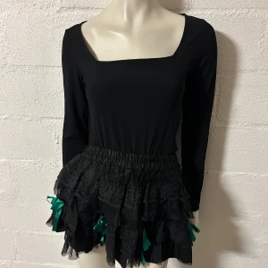 Svart volangkjol med gröna rosetter i onesize - Supersöt svart kortkjol med flera lager volanger och spetsdetaljer. Kjolen har gröna satinrosetter som ger en unik och lekfull look. Perfekt för dig som gillar statement-plagg och vill sticka ut.