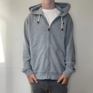 Grå hoodie från Morris Company - Säljer en grå hoodie från Morris Company i storlek M. Tröjan har huva med vita snören och bruna detaljer, dragkedja framtill och två fickor. Liten broderad logga på bröstet. Perfekt för chill dagar och enkel att matcha med jeans eller joggers.