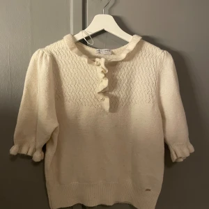 Vit stickad tröja med volang Holly & Whyte - Supersöt vit stickad tröja från Holly & Whyte med volang och pärldetaljer vid halsen. Tröjan har ett diskret mönster upptill, puffade trekvartsärmar och ribbade muddar. Perfekt för dig som gillar romantiska detaljer och mjuka material.