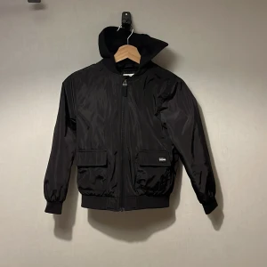 Svart bomberjacka från KappAhl - Svart bomberjacka från KappAhl med huva och dragkedja framtill. Jackan har två fickor med lock och ribbade muddar vid ärmslut och nederkant. Tillverkad i syntetmaterial och har en stilren look som passar till många outfits.