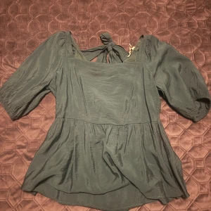 Grön blus med puffärm från AWARE - Säljer en mörkgrön blus från AWARE i storlek S. Blusen har puffiga trekvartsärmar, fyrkantig halsringning och snygga knytband i ryggen som ger en öppen rygg-look. Materialet är mjukt och har en lätt glans. Perfekt för dig som gillar trendiga detaljer och volym.
