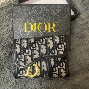 Plånbok - Snygg plånbok med klassiskt monogrammönster i svart och beige. Plånboken har guldfärgad D-detalj framtill, flera fack för kort och ID, samt svart skinn på insidan. Helt oanvänd och redo att skickas! 