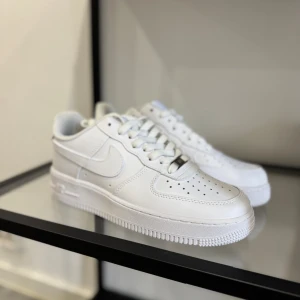 Nike Air Force 1 helvita sneakers - Klassiska Nike Air Force 1 sneakers i helvitt skinn med perforerad tå, rund tå och platt sula. Snygg och clean design med vita snören och ikoniska swoosh-loggan på sidan. Perfekta för dig som vill ha en tidlös och fräsch look.