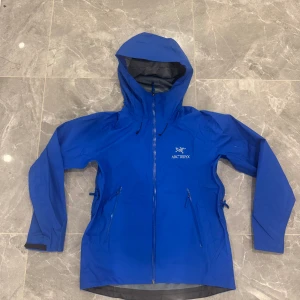 Blå Arc'teryx Gore-Tex vindjacka - Snygg blå vindjacka från Arc'teryx med huva och vattentätt Gore-Tex-material. Jackan har dragkedja framtill, två sidofickor med dragkedja och coolt Arc'teryx-logo på bröstet. Perfekt för dig som gillar outdoor och vill ha en jacka med grym passform och funktion.