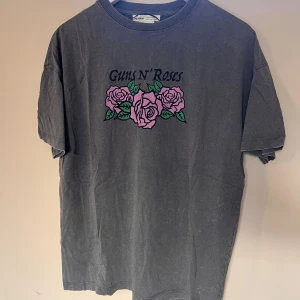 Guns N' Roses t-shirt från Pull&Bear -  Grå t-shirt från Pull&Bear med Guns N' Roses-tryck och stora rosa rosor både fram och bak. Oversized fit med korta ärmar och rund hals. Perfekt för dig som gillar bandmerch och vill sticka ut med en snygg grafisk design.