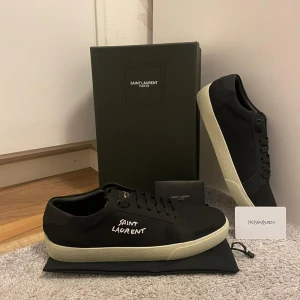 Svarta sneakers från Saint Laurent - Svarta sneakers från Saint Laurent med vit broderad logga på sidan och vit platt sula. Skorna har klassisk snörning och är tillverkade i canvas med detaljer i skinn. Snygg minimalistisk design som passar till allt.
