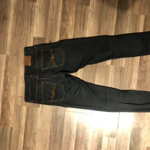 Mörkblå jeans från Nudie Jeans 32/33 - Säljer ett par mörkblå jeans från Nudie Jeans med klassiska orange sömmar och snygga bakfickor med vågig detalj. Jeansen har rak passform och är tillverkade i slitstarkt denimtyg. Perfekta för dig som gillar stilrena och tidlösa jeans.