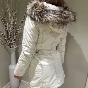 Vit parkajacka med fuskpäls - Snygg vit parkajacka med huva och fluffig fuskpäls runt kanten. Jackan har svarta kontrastsömmar, dragkedja framtill, flera fickor med lock och knapp, samt detaljer vid midjan för extra passform. Perfekt för kalla dagar och riktigt stilren look.
