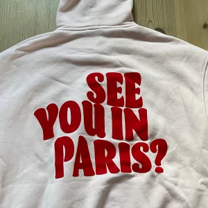 Ljusrosa hoodie med röd text - Ljusrosa hoodie med stor röd text 'SEE YOU IN PARIS?' på ryggen. Tröjan har huva, lång ärm och en klassisk magficka framtill. Tyvärr har den bläck frammtill. Men annars helt ny. 
