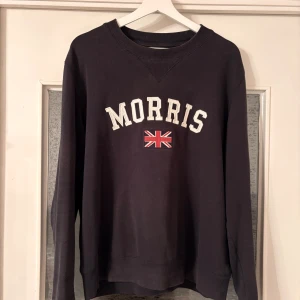 Svart sweatshirt från Morris med tryck - Svart sweatshirt från Morris i storlek small med klassiskt tryck på bröstet och brittisk flagga. Tröjan har rund hals, ribbade muddar och lång ärm. Perfekt för dig som gillar stilren design med en touch av brittisk vibe.