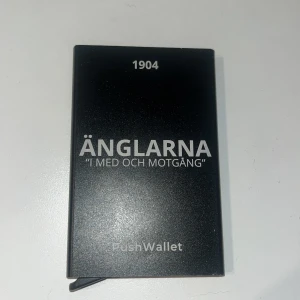 Svart PushWallet plånbok Änglarna - Slimmad svart plånbok i metall med texten 'ÄNGLARNA I MED OCH MOTGÅNG' och årtalet 1904 på framsidan. Plånboken har en minimalistisk design och är perfekt för kort. Snygg och stilren att ha med sig i fickan. Den rymmer max 13 kort. Om ni vill diskutera priser så går det super